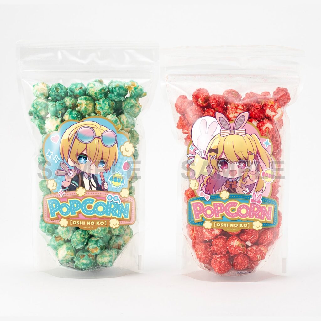 Two popcorn bags side by side. The left bag contains green/blue popcorn and an image of Oshi no Ko character Aqua, whilst the right hand bag contains red popcorn and an image of the character Ruby from Oshi no Ko