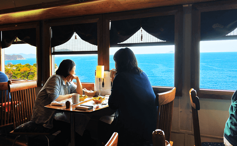 Two people sat at a table on a train, which has good and drink on. They are looking out the window at a view of the sea in Kyoto prefecture.