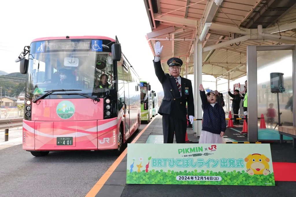 A Pikmin themed bus departing a bus station, being waved off by a man and a child