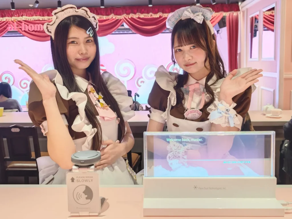 Two maids standing in front of a screen showing an English translation of a Japanese phrase